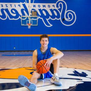 Peyton Mullarkey holds a basketball between his hands as he sits on the Cecil the Sagehen logo in the middle of the basketball court. He has short brown hair and wears a blue practice uniform and gray sneakers. Behind him is a basketball net and a blue wall with Pomona-Pitzer Sagehens in blue text with a white outline. Pomona-Pitzer is in smaller plain script and Sagehens is in large cursive script.